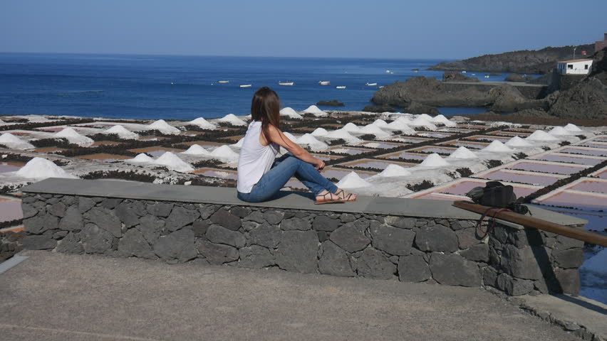 Caucasian woman sitting on a stone wall watching the salt pans and the Atlantic Ocean in La Palma