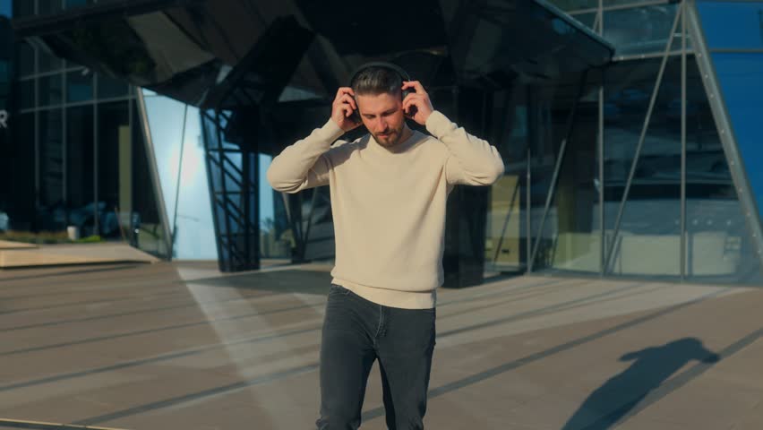 Stylish young man with beard listens to music outdoors, raising arms while grooving to beat. Modern urban lifestyle portrait in casual fashion and relaxed vibe.