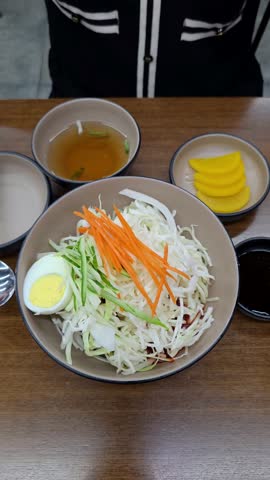 A person uses chopsticks to mix a bowl of Korean Jjolmyeon, combining chewy noodles with fresh shredded cabbage, vegetables, and a spicy gochujang sauce for a traditional and delicious meal