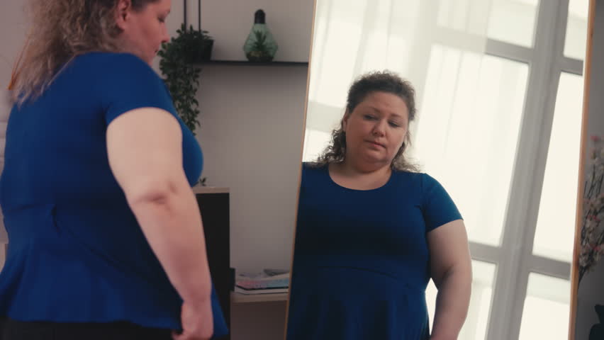 Curvy woman adjusting tight blouse in front of mirror, upset with body changes