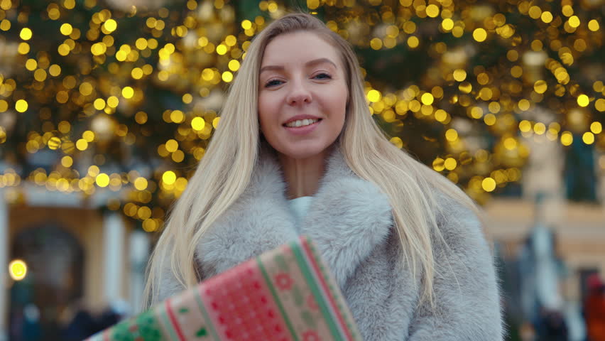 Portrait of smiling woman wearing fur coat holds present near Christmas tree outdoors. Blonde female shows gift box visiting traditional winter fair on central city square
