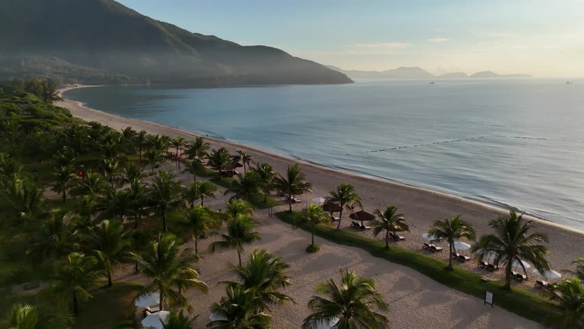 A series of drone clips capturing a beautiful sandy beach at Amiana Resort in Cam Ranh, Vietnam. The footage showcases rows of palm trees, lounge chairs, white umbrellas along the tranquil coastline