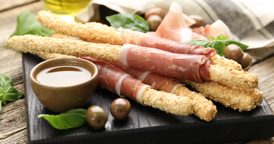 Woman taking grissini breadstick with prosciutto from board at wooden table, closeup
