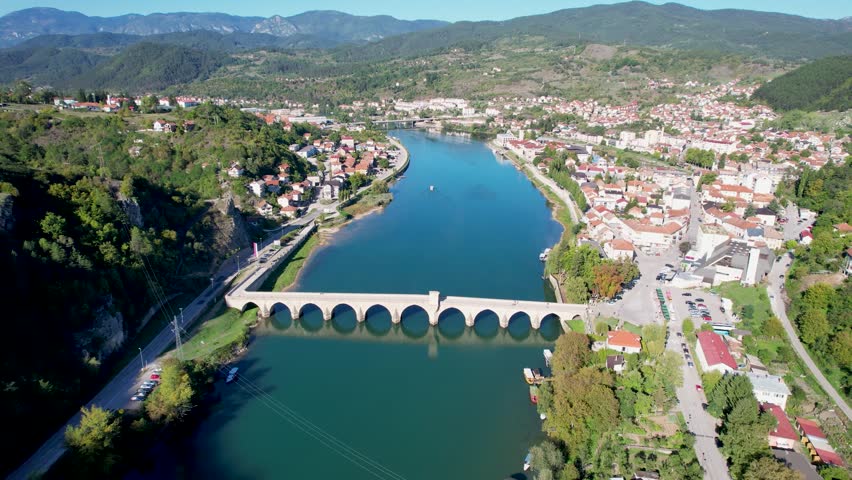Aerial reveal of Mehmed Paša Sokolović Stone Bridge in Visegrad, Bosnia and Herzegovina, spanning the Drina River with surrounding town, houses, hills, and lush greenery in a scenic landscape view