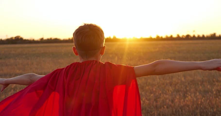 Superhero child silhouette in red cape runs toward sunrise outdoors with arms outstretched as if flying. child freedom, imagination, motivation, and aspiration in open landscape with dramatic sunlight