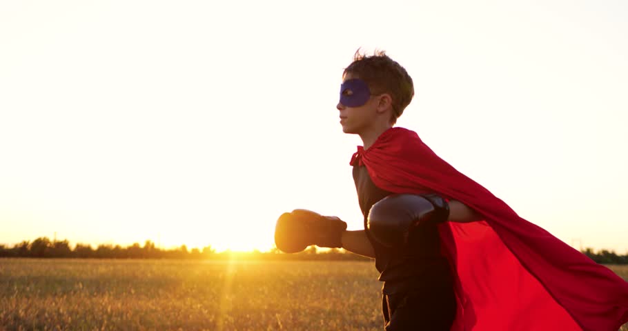 superhero kid runs in meadow during golden hour wearing boxing gloves and red cape. child shows imagination, strength, and playful energy. cape superhero child embraces dream. child running outdoor
