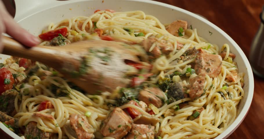 Chef he cook stirs the spaghetti with cherry tomato slices and spinach in creamy sauce and salmon pieces simmering in a pan with dill and green onions, with a wooden spatula.