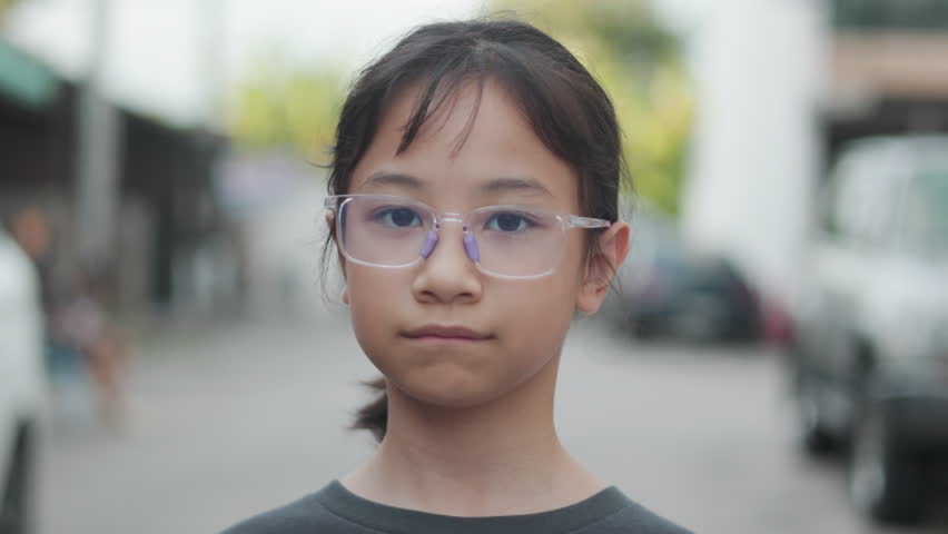 Close up face of adorable Asian girl wearing glasses, looking straight at the camera with a calm expression. Blurred urban background, natural light, capturing childhood innocence and thoughtful mood.