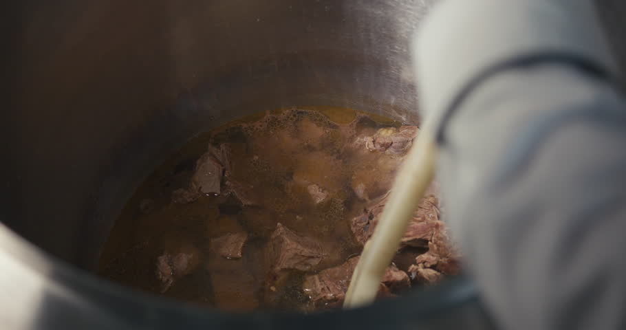 Worker stirring chunks of meat in a large pot with hot liquid, showing the cooking and food preparation process in a professional kitchen.