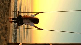 Vertical video of a girl sitting on a swing by the sea at sunrise or sunset, enjoying the peaceful view of the ocean and golden sky. Relaxation, freedom, and summer vacation mood. - Powered by Shutterstock - Get 15% off with code: PIKWIZARD15