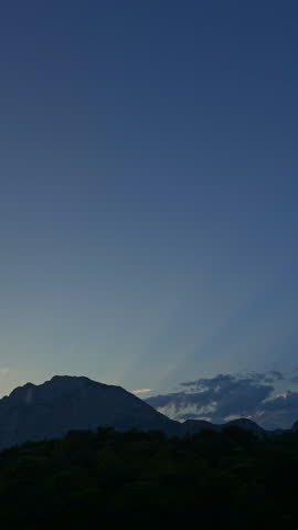 Mountain landscape with sun rays shining from behind the peaks, few clouds in the sky, warm summer atmosphere at sunrise or sunset, peaceful and scenic view.