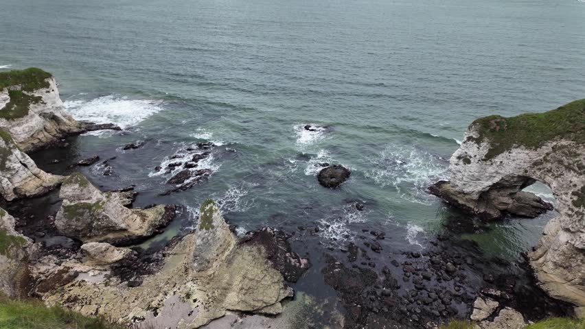 A dramatic cliffside seascape with waves breaking against rocks and a striking natural geological arch along the Atlantic Ocean and rugged coastline of Northern Ireland