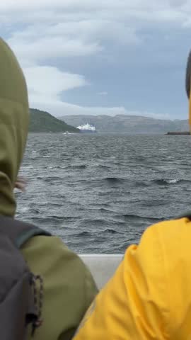 Two people in colorful jackets watch the stormy waters of the Barents Sea from a small boat, with a large vessel visible on the horizon