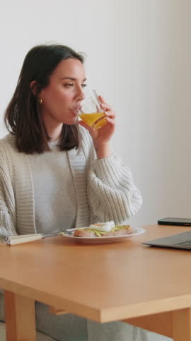 Vertical video, A young woman enjoys a healthy breakfast of avocado toast and orange juice while working remotely at her dining table.
