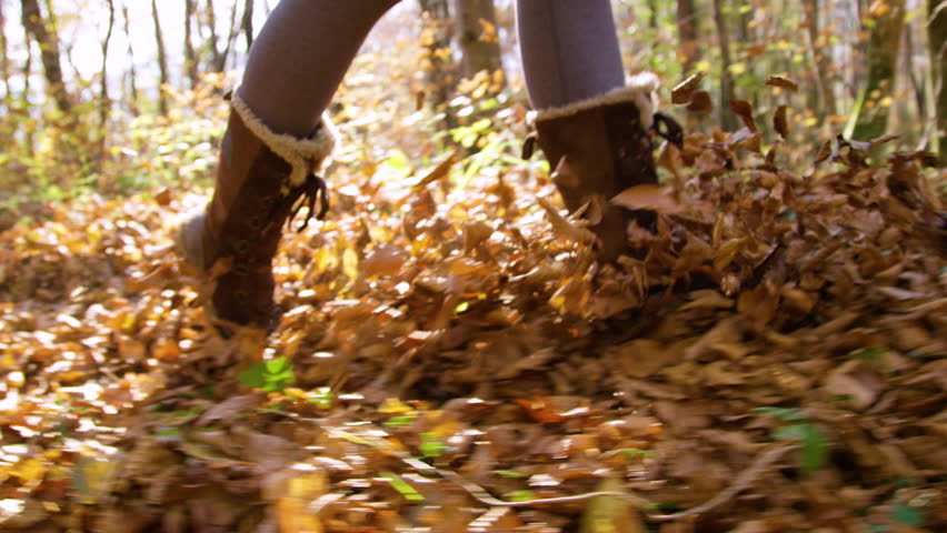 LENS FLARE, SLOW MOTION, CLOSE UP: Fallen autumn leaves fly through the air as woman in warm boots runs along a forest path. Playful adult enjoys the beauty of nature in fall season on a sunny day.