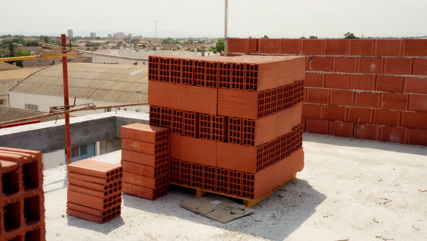 Hollow red clay bricks stacked on a wooden pallet at a sunny construction site, with a newly built wall and distant city skyline under a clear blue sky, ready for masonry work