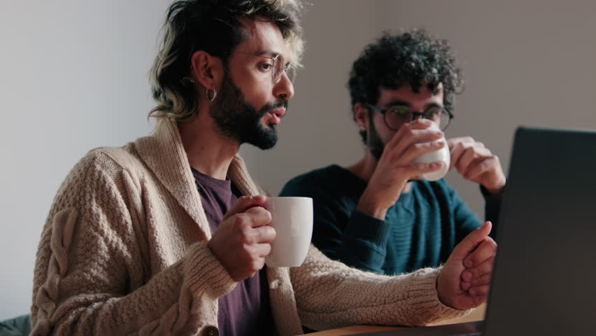 A gay couple sits together at their home office, drinking coffee and working on their laptop. They discuss business and look happy.