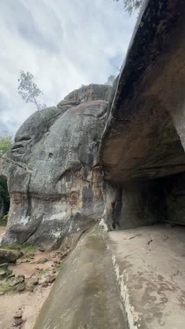 A unique rock formation with a large overhang creates a natural cave, with mossy textures and ancient carvings visible on the stone walls under a cloudy sky