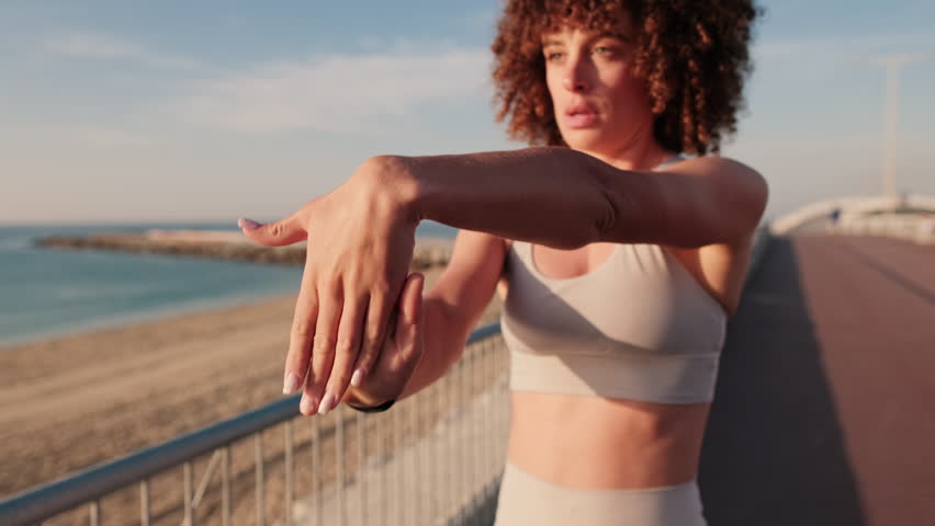 Young woman stretching her wrist by the beach at sunrise. Getting ready for a workout by the sea. Health, fitness, and active lifestyle.