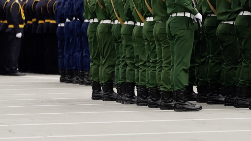 Disciplined military formation with uniformed personnel in green and dark dress standing in precise ranks on a marked concrete parade ground during a ceremony, boots aligned.