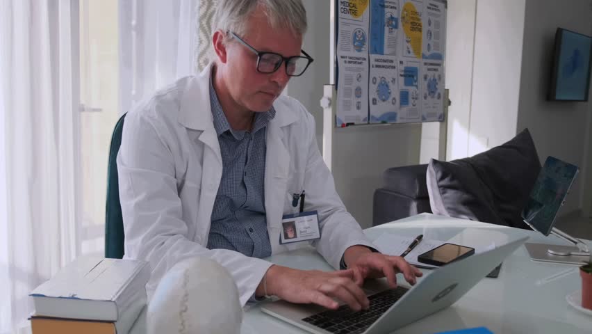 Focused senior doctor in glasses and white coat typing on laptop in modern medical office. Healthcare professional working with documents and digital records at desk. - Powered by Shutterstock - Get 15% off with code: PIKWIZARD15