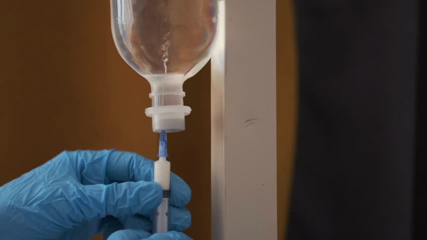 Gloved medical worker adjusting IV drip and filling syringe from infusion bottle. Close-up, nurse hands performing intravenous procedure in hospital environment. Professional healthcare treatment.