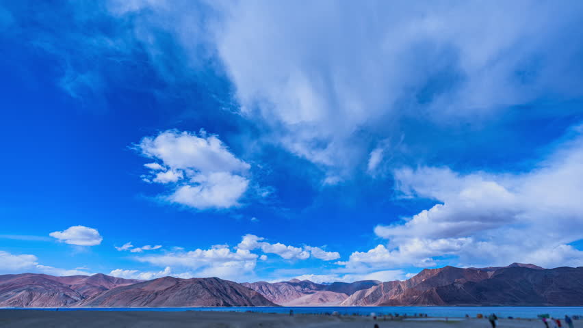 Timelapse A wide-angle landscape photography showcasing Pangong Lake with a backdrop of rugged Himalayan mountains under a vibrant, cloud-filled sky. Tourists are scattered along the lakeshore.