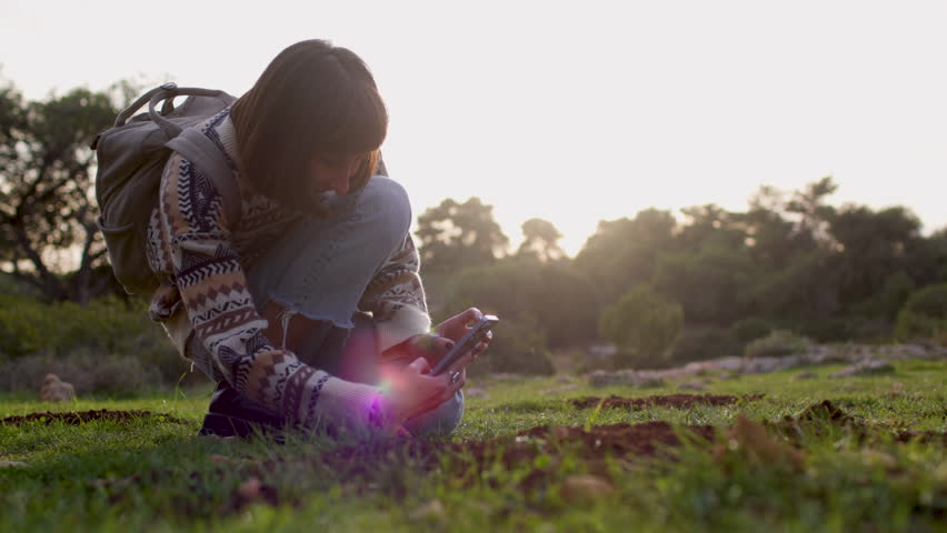 A side view of a girl photographing a beautiful plant during a hike. A young brunette with a backpack touches her phone screen, taking a photo of a flower. Modern technology, nature concept.