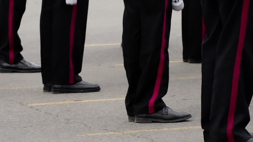 Close-up of uniformed personnel standing in precise formation on asphalt, black trousers with red stripes and polished shoes visible during a formal drill or parade lineup.