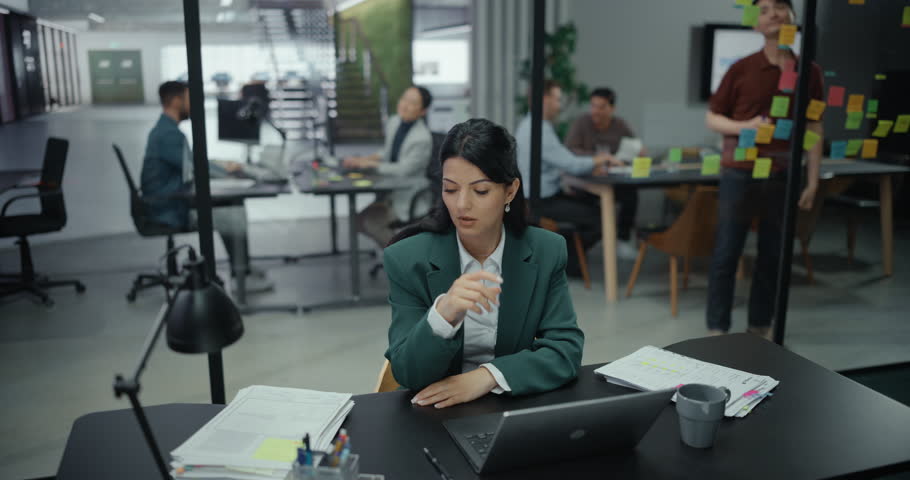 Multiethnic Female Employee Trying to Focus on a Task, Feeling Under the Weather, Ill and Unwell while Writing a Report on a Laptop Computer, Sitting Behind a Desk in a Modern Office