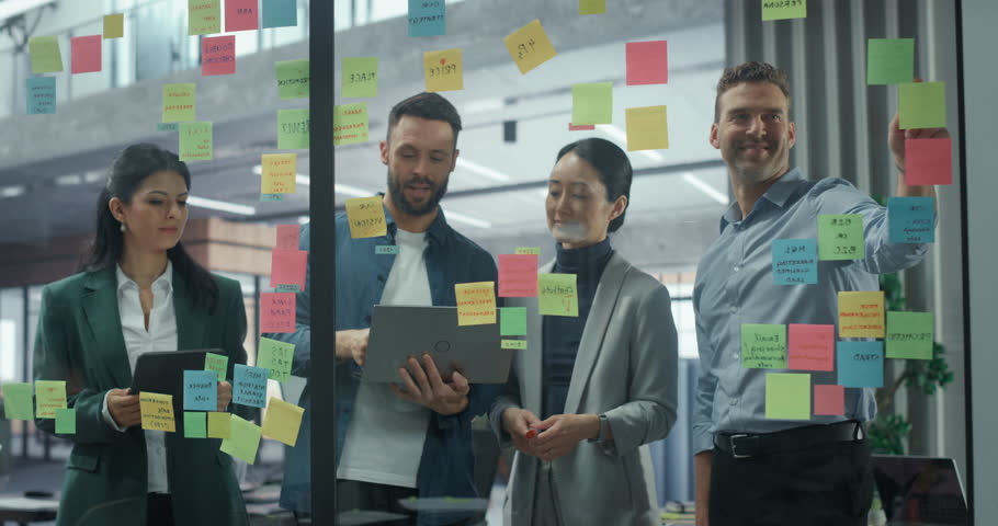 Employees Engage in a Creative Planning Session, Writing On and Placing Post-It notes on a Glass Wall to Structure a Business Project. They Discuss Each Suggestion and Rearrange Notes