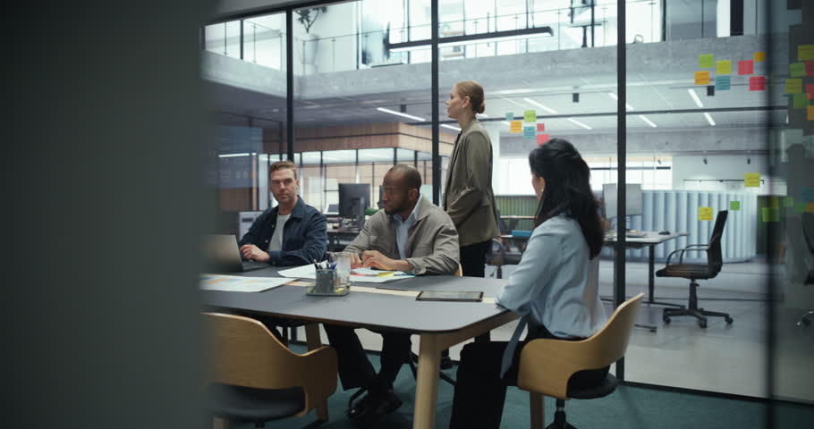 Female Business Leader Stands at the Front of a Conference Room, Delivering a Presentation with Charts and Financial Data on Screen. Colleagues Sit Around the Table, Listening and Taking Notes
