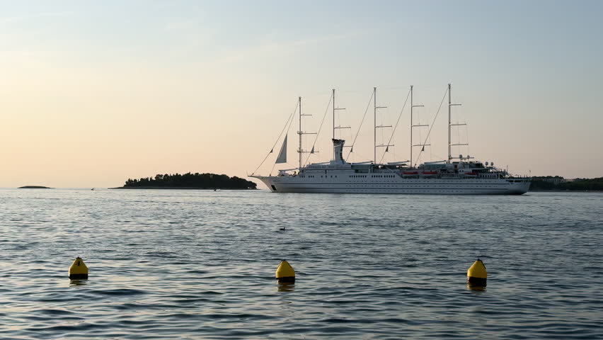 A luxury cruise ship sails past Rovinj, Croatia during a serene sunset. Calm Adriatic waters reflect soft golden light and silhouettes of the coastline.