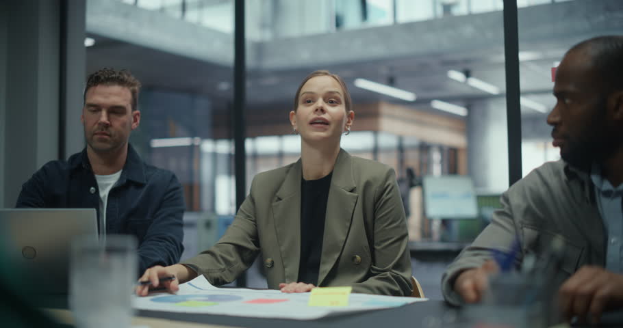 In a Corporate Boardroom, Woman Speaks Confidently to Coworkers Sitting Around a Table. Employees Follow Her Presentation, Discussing Strategies and Analyzing Company Reports