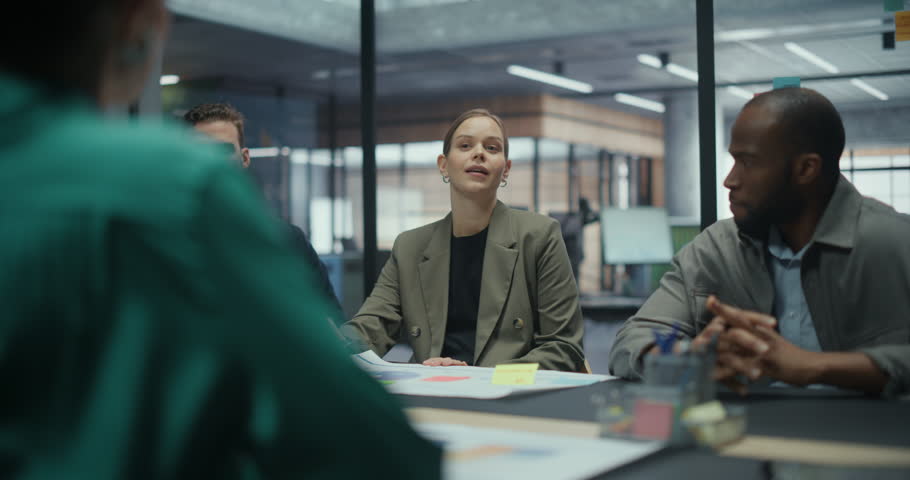 Female Manager Leads a Business Meeting in a Modern Office Conference Room, Addressing Her Colleagues Across the Table. Team Members Listen and Review Documents as She Explains Project Details