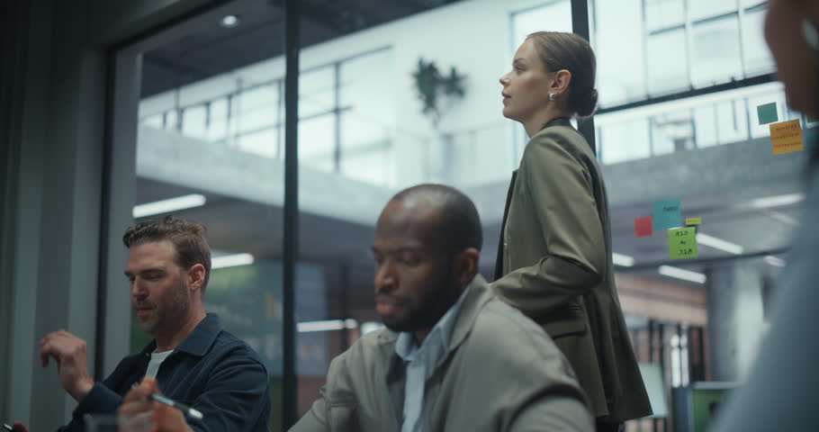 Woman Leads a Corporate Presentation in a Meeting Room, Pointing to Charts and Diagrams on a Large Display. Employees Listen Carefully, Considering Her Ideas for Future Projects and Company Goals
