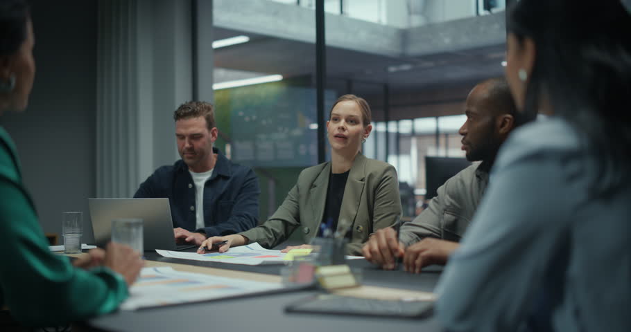 Female Executive Talking with Her Team in a Meeting, Sitting with Colleagues Behind a Table Filled with Computers and Papers. She Outlines Key Objectives while the Group Listens