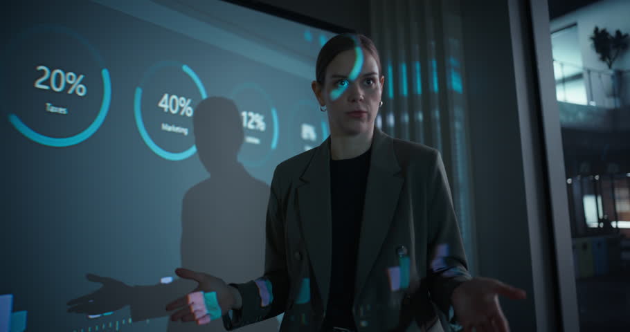 In a Dark Office Space, Female Manager Stands at the Front of the Room Delivering a Corporate Presentation. She Outlines Project Objectives to the Team while Data and Visuals Appear on Screen