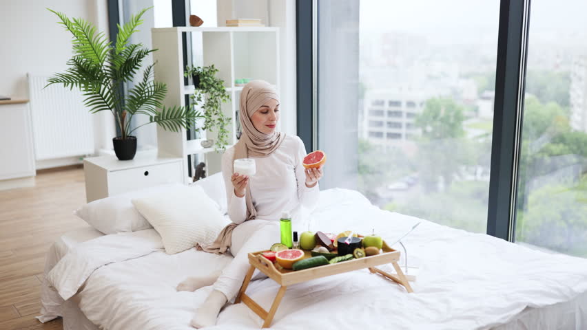 Young Muslim woman in her 20s sitting indoors preparing to use grapefruit and cream as part of her skincare routine, promoting natural wellness and self-care.
