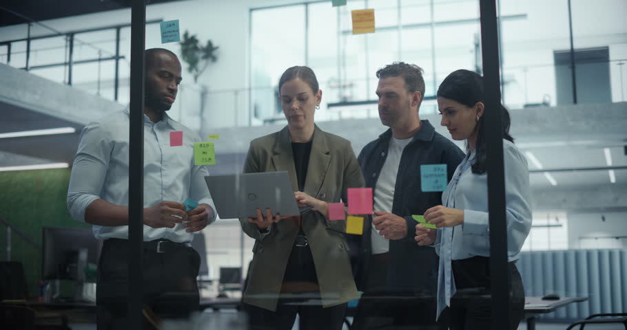 Employees Engage in a Creative Planning Session, Writing On and Placing Post-It notes on a Glass Wall to Structure a Business Project. They Discuss Each Suggestion and Rearrange Notes