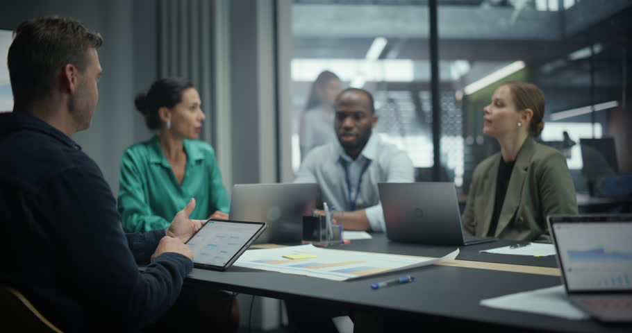 Team of Executives Talking About Business in a Meeting, Sitting Together Behind a Table Filled with Computers and Papers. Colleagues Outline Key Objectives while the Group Listens