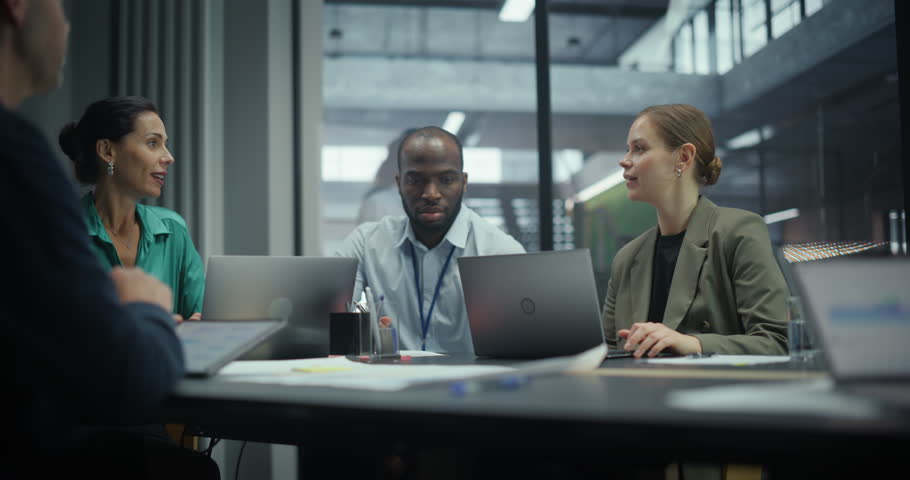Employees Sitting Side by Side in a Meeting in Conference Room, Working on Laptop Computers and Having a Conversation. Group is Busy with Reports and Project Planning for a Corporate Client