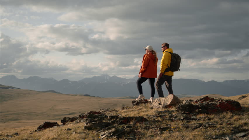 A couple of tourists hikers, a man and a woman, stand on a mountaintop, hugging and joyfully raising their hands. The young people are enjoying the beautiful mountain scenery and outdoor activities.