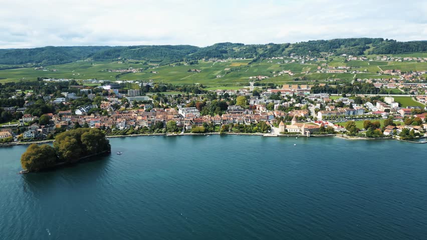Aerial view of Place des Tilleuls where red-roofed buildings meet the blue waters of the lake, creating a stunning contrast against the backdrop of lush green hills, Rolle, Vaud, Switzerland.