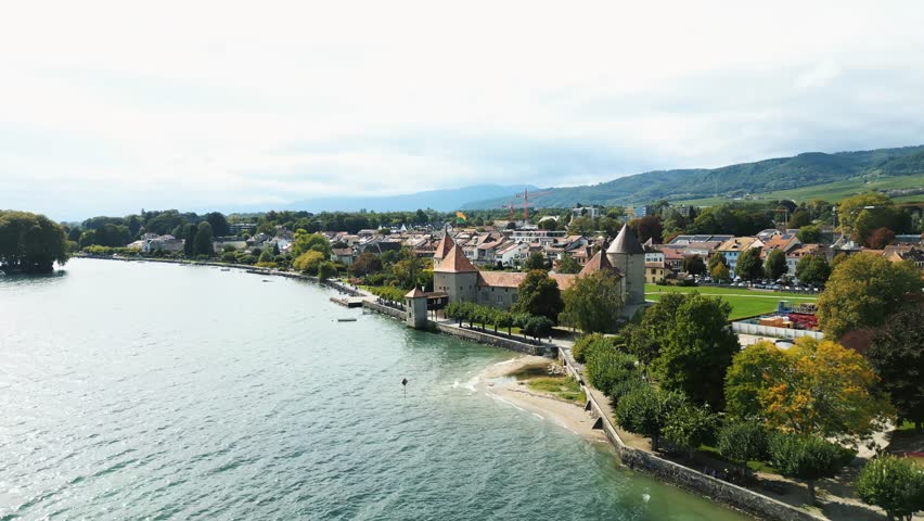 Aerial view of the medieval Rolle Castle with its red rooftops, standing proudly beside the serene Lake Geneva and lush greenery, Rolle, Vaud, Switzerland.
