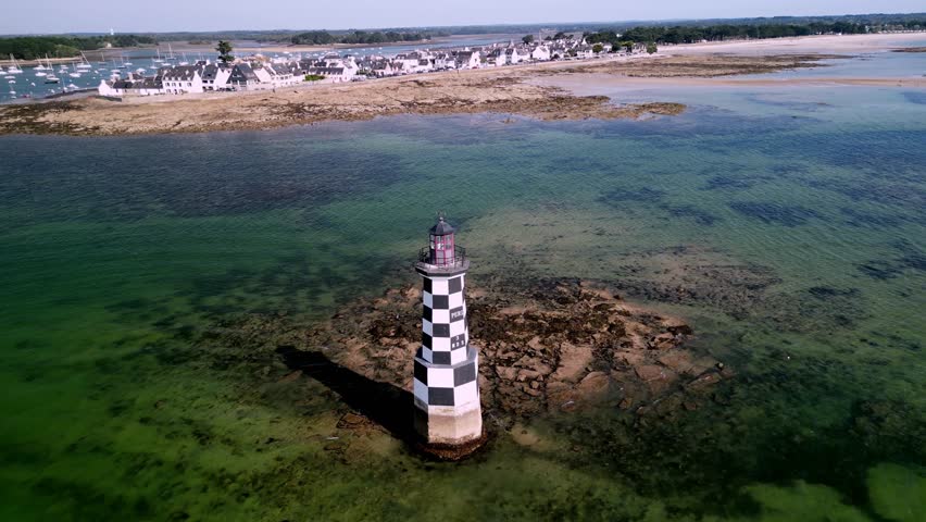 Aerial view of the striking black and white checkered Phare de la Perdrix lighthouse, surrounded by shimmering turquoise waters and rocky shores, Île-Tudy, Brittany, France.