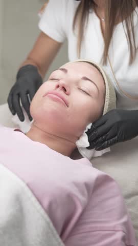 A woman is receiving a professional facial cleansing treatment at a beauty salon. The cosmetologist, wearing gloves, gently wipes the clients face with soft tissues, completing the skincare procedure