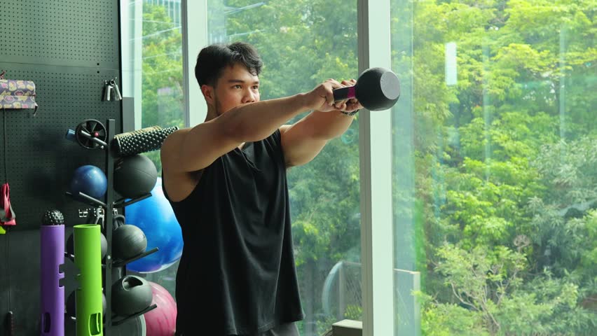 Athletic man performs kettlebell swing in sunlit gym with modern equipment and large windows.