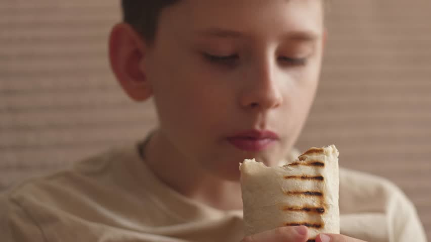 A portrait of a Caucasian boy, 11-12 years old, eating a delicious burrito while inside, the teenager eats fast food