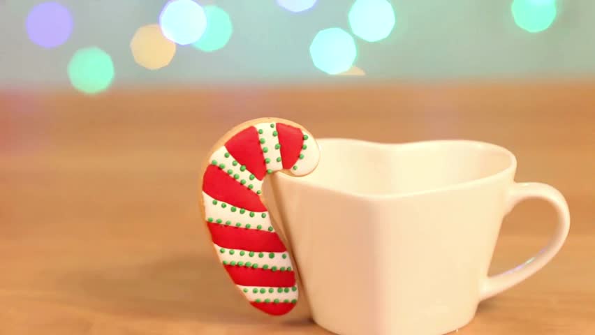 A beautifully decorated candy cane cookie rests on a white heart shaped mug. Festive bokeh lights twinkle in the background creating a warm, cozy holiday atmosphere.